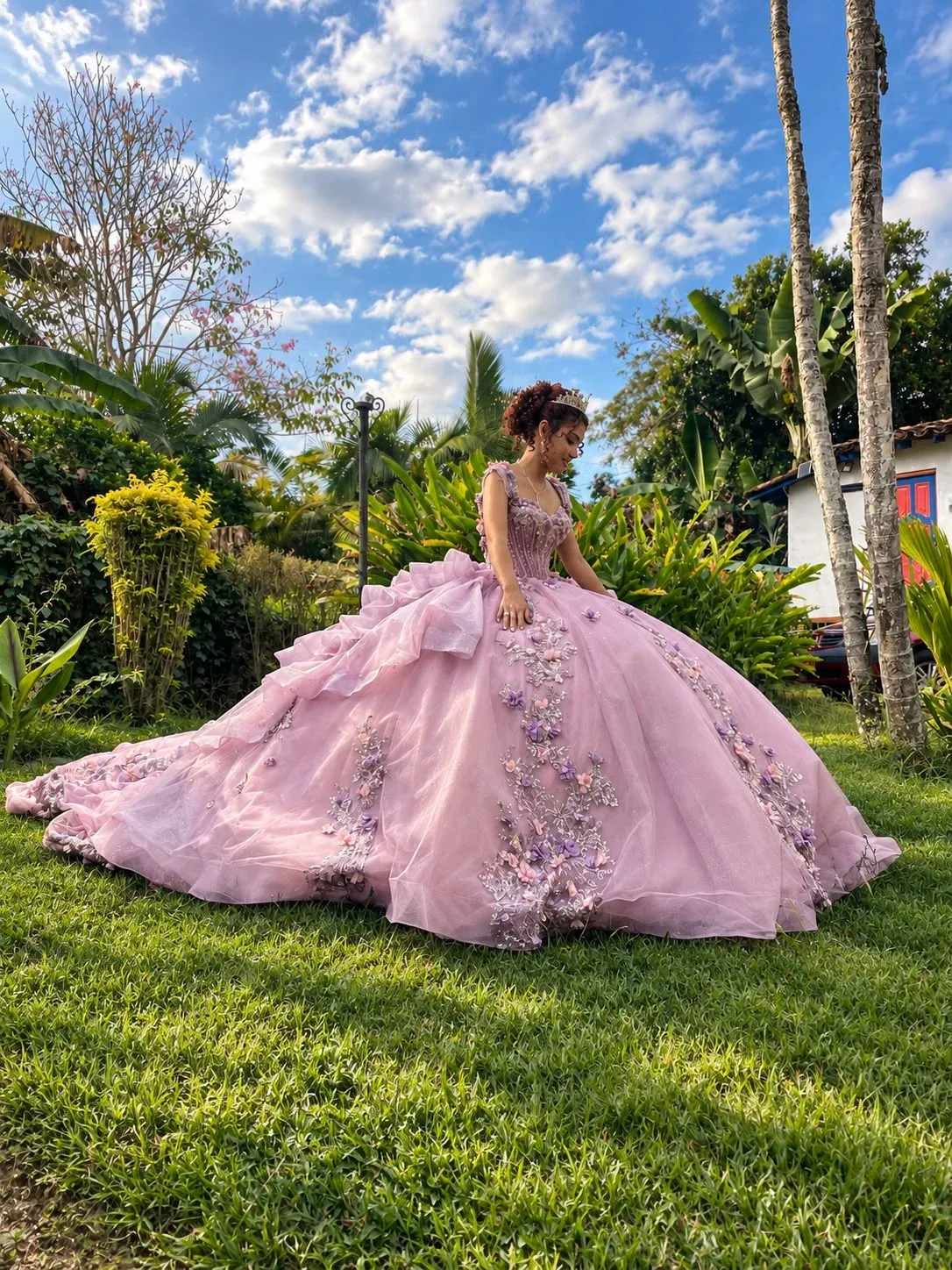 Quinceañera con peinado recogido y vestido rosa lila con flores aplicadas, sentada sobre el césped de un jardín.